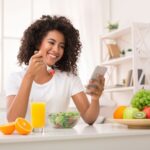 Dieta para gordura no fígado: mulher sorridente com celular na mão comendo salada, com frutas e suco de laranja na mesa.