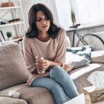 Dispepsia, o que é: mulher sentada no sofá, encolhida, com dor no abdômen.