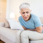 o que é constipação​: mulher sentada no sofá com dor abdominal